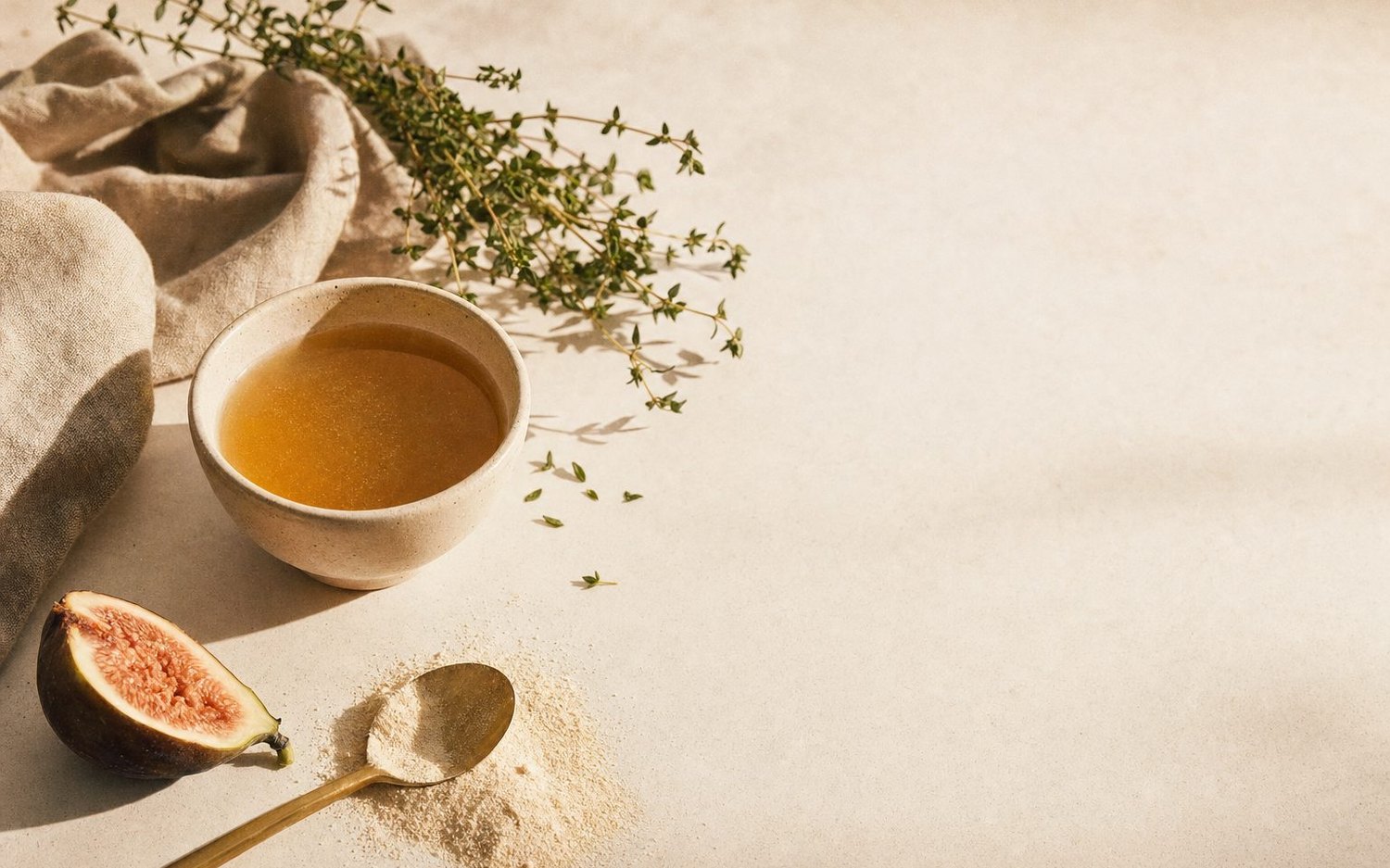 A ceramic bowl of golden broth with fresh herbs, a fig and collagen powder on warm ivory linen